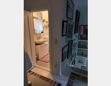 Ein kleiner Flur mit gerahmten Bildern an einer Wand führt in ein Badezimmer mit weißen Fliesen, weißem Waschtisch und Schrank sowie einem beleuchteten Spiegel. Der Boden des Badezimmers ist mit einem rosa Teppich bedeckt.