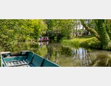 Eine ruhige Flusslandschaft mit dichtem, grünen Uferbewuchs; im Vordergrund ein leeres grünes Boot, im Hintergrund eine Gruppe von Menschen in einem Kahn, der von einem Fährmann mit einer Stange bewegt wird, und eine kleine Holzbrücke über dem Wasser.