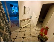 Eingangsbereich einer Wohnung mit hellem Teppich und schwarz-weißem Rautenmuster. An der Wand stehen eine schmale Kommode, ein Spiegel und ein Bild, daneben führt eine Tür ins Badezimmer; ein weiterer Durchgang zeigt einen Wohnraum mit blauem Licht und einem roten Teppich mit Kreisdesign.