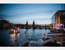 Abendliche Flusslandschaft mit der beleuchteten Oberbaumbrücke im Hintergrund, Booten auf dem Wasser und modernen Gebäuden sowie einer Veranstaltungsbühne am Ufer im Vordergrund.