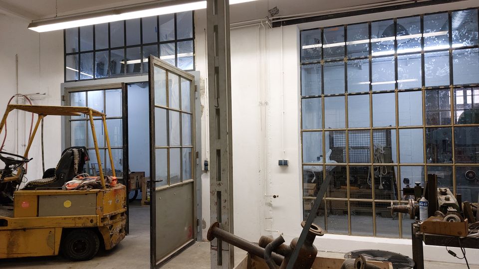 Industriehalle mit weiß gestrichenen Wänden und großen Fenstern mit Metallrahmen. Im Vordergrund steht ein gelber Gabelstapler, und im Hintergrund sind Werkzeuge und Maschinen sichtbar.