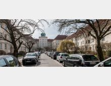 Straßenansicht einer Wohnsiedlung mit symmetrisch angeordneten Altbaugebäuden und Satteldächern. Entlang der Straße stehen geparkte Autos, während kahle Bäume und ein einzelner Weidenbaum das Straßenbild prägen; im Hintergrund ist ein mehrstöckiges Gebäude mit einem Turm zu sehen.