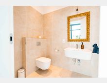 Modernes Badezimmer mit beigen Wand- und Bodenfliesen, einem wandhängenden WC, einem Waschbecken mit brauner Seifenspenderflasche und einem goldgerahmten Spiegel. Ein kleines Fenster mit weißem Rahmen ist ebenfalls sichtbar.