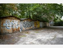 Reihe alter, rostiger Garagen mit Graffiti an den Türen, umgeben von Bäumen und dichtem Grün in einem verlassenen Hof. Der Boden ist mit Beton und einzelnen Blättern bedeckt.