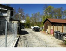 Gepflasterte Gasse mit parkenden Autos und einem Radfahrer im Hintergrund, umgeben von einfachen Gebäuden aus Holz und Stein. Die Szene wird von Bäumen und blauem Himmel eingerahmt, mit Mülltonnen und Zäunen am Straßenrand.