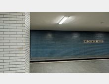 U-Bahn-Station mit türkisfarbenen Fliesenwänden, einer weißen Decke mit rechteckiger Leuchtstofflampe und einem Schild mit der Aufschrift „Parchimer Allee“. Im Vordergrund ist eine Säule mit weißen Kacheln und eine geflieste Bahnsteigfläche zu sehen.