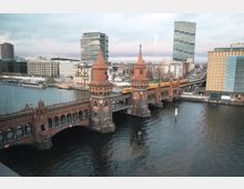 Ansicht der Oberbaumbrücke in Berlin, einer roten Ziegelbrücke mit zwei Türmen über der Spree, im Hintergrund moderne Bürogebäude und Hochhäuser. Eine U-Bahn fährt auf der oberen Ebene der Brücke, während das Wasser der Spree im Vordergrund zu sehen ist.