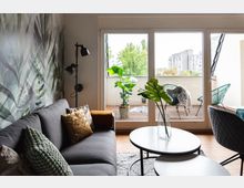 Wohnzimmer mit einer grauen Couch und dekorativen Kissen vor einer Wand mit Pflanzen-Muster. Große Fenster führen zu einem Balkon mit Holzboden, grünen Pflanzen und Sitzmöbeln, im Hintergrund sind Bäume und Wohnhäuser sichtbar.