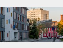 Straßenszene mit einem modernen Gebäude auf der linken Seite, das durch bunte Fensterrahmen gekennzeichnet ist, und einem roten historischen Gebäude rechts. Im Hintergrund ist ein großes Hochhaus mit der Aufschrift „Europa Universität“ erkennbar.