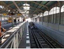 Überdachte U-Bahn-Station mit zwei Bahnsteigen, Schienen in der Mitte und gelben Zügen auf der linken Seite. Die Halle hat ein gewölbtes Dach mit Stahlträgern und große Fenster auf beiden Seiten, durch die Tageslicht einfällt.