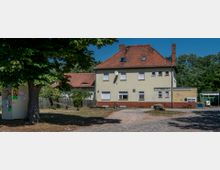 Zweistöckiges, beiges Gebäude mit rotem Ziegeldach und Satellitenschüssel, umgeben von Bäumen und Rasenflächen. Im Vordergrund ein gepflasterter Weg, ein großer Baum und ein Altglascontainer; rechts ein Biergarten-Eingang.