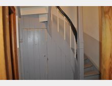 Eine enge Innentreppe aus Holz mit abgenutztem grauen Anstrich und schwarzem, glänzend lackiertem Handlauf. Rechts von der Treppe befindet sich eine Wand, links ein graues Holztür-Panel mit sichtbaren Gebrauchsspuren.