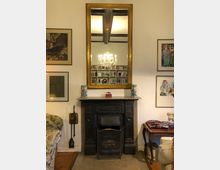 Wohnzimmer mit einem antiken Kamin und einem großen, goldgerahmten Spiegel darüber. Der Spiegel reflektiert einen Kronleuchter und Bücherregale im Hintergrund. Dekorationen umfassen Wandbilder, Vasen und Möbel wie einen Beistelltisch und eine Polstercouch.