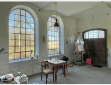 Innenraum eines Ateliers mit hohen, bogenförmigen Fenstern und Blick auf eine Außenlandschaft. Der Raum ist schlicht gestaltet mit einem Holztisch, Stühlen, Malutensilien, einer Staffelei, und einer rustikalen Holztür im Hintergrund.