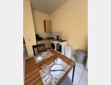 Kleine Küchenzeile in einem hellen Raum mit Holzschränken, Spüle, Waschmaschine und freistehendem Herd vor einer gefliesten Rückwand. Im Vordergrund steht ein Esstisch mit gedeckten Plätzen; daneben sind zwei Mülleimer auf dem Fliesenboden.