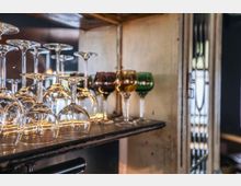 Glasregal mit auf den Kopf gestellten durchsichtigen Weingläsern und farbigen Kelchgläsern, in einem rustikalen, leicht beleuchteten Innenraum.