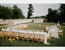 Lange, festlich gedeckte Tafeln mit weißen Tischdecken, Glasgeschirr und Holzstühlen stehen auf einer Rasenfläche im Freien. Im Hintergrund verläuft eine weiße Balustrade entlang einer Terrasse mit Bäumen und einem Wasserlauf daneben.