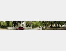 Straßenszene mit einem weißen Wohnhaus, das von Bäumen und einem weißen Holzzaun umgeben ist, sowie einem geparkten roten Fahrzeug im Vordergrund. Im Hintergrund rechts ein kleiner Parkbereich mit Bäumen und offenen Flächen, daneben Pflasterstraße und Verkehrsschild.