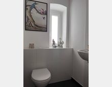 Kleines Badezimmer mit weiß gefliesten Wänden, einer Toilette links und einem kleinen Waschbecken rechts. Über der Toilette hängt ein großes Bild mit Vogelmotiven, und auf der Fensterbank stehen Dekofiguren vor einem schmalen, weißen Fenster.