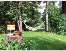 Ein Garten mit grüner Wiese, umgeben von Bäumen und Sträuchern. Im Vordergrund steht ein alter Baumstumpf mit einer orange-weiß gestreiften Tischlampe und einem Blumentopf, im Hintergrund hängt eine helle Hängematte zwischen zwei Bäumen.