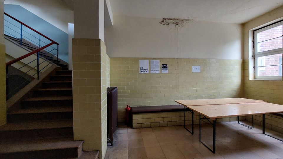 Ein einfacher Innenraum mit gelb-beigefarbenen Fliesenwänden, einem Holztisch mit Metallgestell und einer Bank an der Wand. Links führt eine Treppe mit rotem Geländer nach oben, und rechts befindet sich ein Fenster mit sichtbaren Außenziegelmauern.