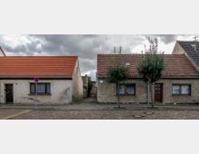 Straßenszene mit zwei einstöckigen Wohnhäusern, die braune Dachziegel und einfache Fassaden haben; dazwischen ist ein schmaler Durchgang sichtbar. Vor den Häusern stehen junge Bäume auf einem gepflasterten Gehweg, und die Straße ist mit Kopfsteinpflaster bedeckt.