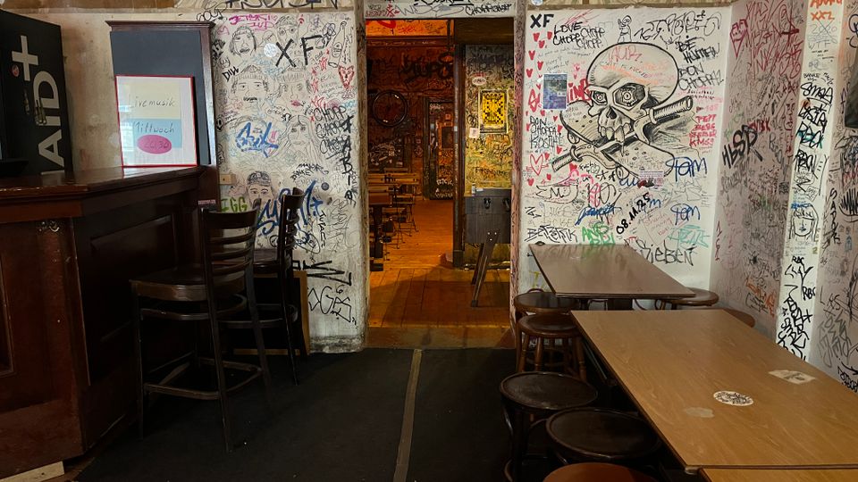 Innenraum einer alternativen Kneipe mit eng gestellten Holztischen und Hockern sowie einer dunklen Theke links. Die Wände und der Durchgang in den hinteren Raum sind dicht mit Graffiti und Zeichnungen bedeckt, darunter ein großes Totenkopf-Motiv.