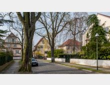 Straßenszene in einem Wohngebiet mit mehreren Häusern im Stil der 1920er Jahre, roten Ziegeldächern und hellen Fassaden. Die Straße ist gepflastert, gesäumt von Bäumen und Gehwegen, mit parkendem Auto und Grünanlagen im Vordergrund.