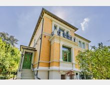 Ansicht einer mehrstöckigen Gründerzeit-Villa aus gelbem Backstein mit weißen Stuckelementen, grün gerahmten Fenstern und kleinem Balkon mit schmiedeeisernem Geländer. Links führt eine Treppe zu einer grünen Eingangstür, umgeben von Gartenbäumen und Sträuchern.