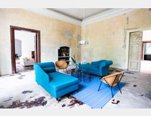 Wohnzimmer in einem Altbau mit abgenutzten Wänden und Boden. Die Einrichtung besteht aus blauen Polstermöbeln, modernen kleinen Tischen, einem Teppich in Blau und Orange, sowie einem Ledersessel; im Hintergrund sind eine offene Tür und ein Kamin sichtbar.