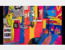 Ein lebhaft gestalteter Innenraum mit bunten, neonfarbenen Wänden in Gelb, Pink und Blau, die mit abstrakten Gemälden, Skizzen und Kunstobjekten dekoriert sind. Eine Regalablage mit Farbdosen und künstlerischen Materialien steht in der Ecke.