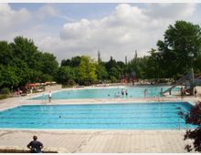 Freibad mit mehreren Becken, darunter ein großes Schwimmbecken im Vordergrund und ein flaches Freizeitbecken im Hintergrund. Umgeben von grünen Bäumen, einem Spielplatz und einem Bereich mit Sonnenschirmen und Sitzgelegenheiten.