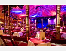 Beleuchteter Innenraum eines Varieté- oder Theatersaals mit runden Tischen, die mit weißen Tischdecken und roten Tischsets dekoriert sind. Kerzenlicht auf den Tischen, samtbezogene Stühle und eine Bühne mit blauem Vorhang sowie eine bunte Zeltdecke mit Lichtprojektionen prägen das Ambiente.