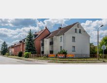 Mehrfamilienhäuser an einer ruhigen Straßenkreuzung, mit einem weißen Gebäude im Vordergrund und einem roten Gebäude dahinter; ein niedriger Zaun mit Werbeplakaten für den „Circus Agros“ befindet sich vor den Häusern. Im Hintergrund sind Bäume und ein klarer Himmel mit Wolken zu sehen.