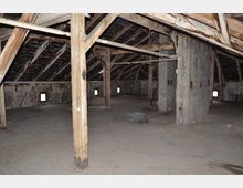 Dachboden eines alten Gebäudes mit sichtbaren Holzstützen, einer schrägen Dachkonstruktion und unverputzten Wänden. Der Raum ist größtenteils leer, mit kleinen Fenstern entlang der Wand und einem Betonboden.