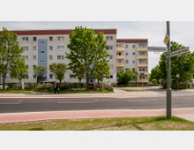 Ein mehrstöckiges Wohngebäude mit heller Fassade, blauen und gelben Akzenten steht hinter einer Straße mit Fußwegen und grünen Rasenflächen. Vor dem Gebäude sind mehrere Bäume, Büsche und parkende Autos zu sehen; Straßenschilder markieren eine Kreuzung.