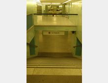 Treppenabgang in einer U-Bahn-Station mit hellgelben und türkisfarbenen Fliesenwänden. Ein Schild mit der Aufschrift "nur Ankunft" hängt oberhalb des Eingangs zur unteren Ebene, die durch Geländer und Beleuchtung abgegrenzt ist.