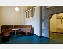 Ein Raum mit einer halbkreisförmigen Holzsitzbank und einem runden Holztisch, beleuchtet durch eine einzelne hängende Kugelleuchte. Die Wand zeigt weiße und gelbe Glaselemente in rechteckigen Rahmen, und rechts steht ein sichtbarer Sandsteinbogen.