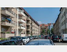 Straßenszene in einer Wohngegend mit mehrstöckigen Gebäuden im Stil der 1920er-Jahre, links mit Balkonen voller Pflanzen und Blumen. Geparkte Autos säumen die Straße, im Hintergrund sind weitere Häuser mit roten Ziegeldächern zu sehen.