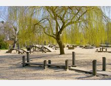 Spielplatz mit Holzelementen und Sandboden, umgeben von einer weinenden Weide mit langen, grünen Zweigen. Im Hintergrund befinden sich weitere Kletterstrukturen und Natursteine.