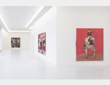 Helle, moderne Kunstgalerie mit weißen Wänden und klaren Linien. An den Wänden hängen großformatige, abstrakte Gemälde mit figürlichen Darstellungen, darunter ein Bild mit rotem Hintergrund.