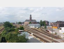 Bahngelände mit mehreren Gleisen, umgeben von verschiedenen Gebäuden, darunter ein markanter Backsteinbau mit Turm im Hintergrund. Im Vordergrund sind Schrebergärten und kleinere Häuschen zu sehen, neben dem Gleisbereich befinden sich Lagerhallen und weitere Betriebsgebäude.