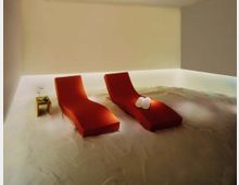 Ein minimalistischer Entspannungsraum mit einem sandbedeckten Boden, zwei roten Liegen und weicher indirekter Beleuchtung. Ein kleiner Holzbeistelltisch mit einem Getränk steht neben einer der Liegen.