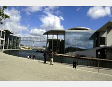 Modernes Gebäudekomplex am Ufer eines Kanals mit breitem, gepflastertem Vorplatz und einer Fußgängerbrücke im Hintergrund. Große Glasfassaden und markante runde architektonische Elemente prägen die Fassade.