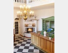 Innenansicht eines hellen Raumes mit einem Kamin, einer hölzernen Theke und einem grünen Vitrinenschrank. Der Raum hat einen schwarz-weiß karierten Boden, eine hängende goldene Kronleuchterlampe und dekorative Details wie Pflanzen und Kerzen.