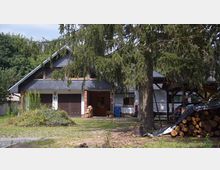 Ein Einfamilienhaus im ländlichen Stil mit grauem Schieferdach und einer dunklen Doppelgarage. Im Vordergrund stehen ein großer Nadelbaum, ein Stapel Brennholz und eine teils begrünte Hofeinfahrt.