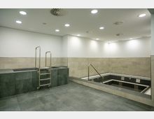 Ein moderner Innenraum mit zwei rechteckigen Becken, eines erhöht mit einer Metallleiter und das andere eingelassen mit einer Metallhandlauf-Treppe. Die Wände sind teils beige gefliest, der Boden aus grauen Fliesen, und die Decke verfügt über runde Einbauleuchten.