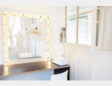 Ein schminkraum mit einem großen beleuchteten Spiegel, darunter ein schwarzer Tisch und ein weißer Stuhl. Auf dem Tisch liegen gefaltete Handtücher, im Hintergrund ein Kleiderständer mit Holzkleiderbügeln und eine Schaufensterpuppe vor einem Fenster mit Vorhang.