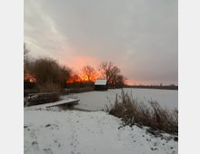 Ein verschneiter See mit einer kleinen hölzernen Hütte am Ufer, umgeben von kahlen Bäumen und Schilf. Im Hintergrund ist ein leuchtender Sonnenuntergang zu sehen, der den Himmel in warmen Rot- und Orangetönen färbt.