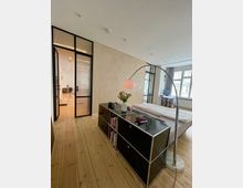 Minimalistisch eingerichtetes Schlafzimmer mit einem Bett, einem schwarzen Regal mit integrierten Fächern, einer gebogenen Stehlampe und hellem Holzboden. Im Hintergrund befinden sich große Fenster und eine Glastür mit schwarzem Metallrahmen, die zu einem weiteren Raum führt.